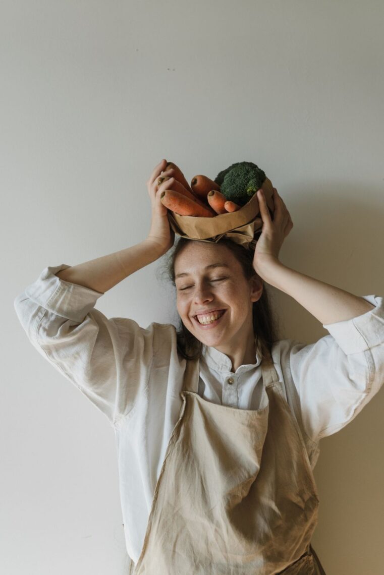 Cheerful woman in apron holding a bowl of vegetables on her head, radiating joy and warmth.
